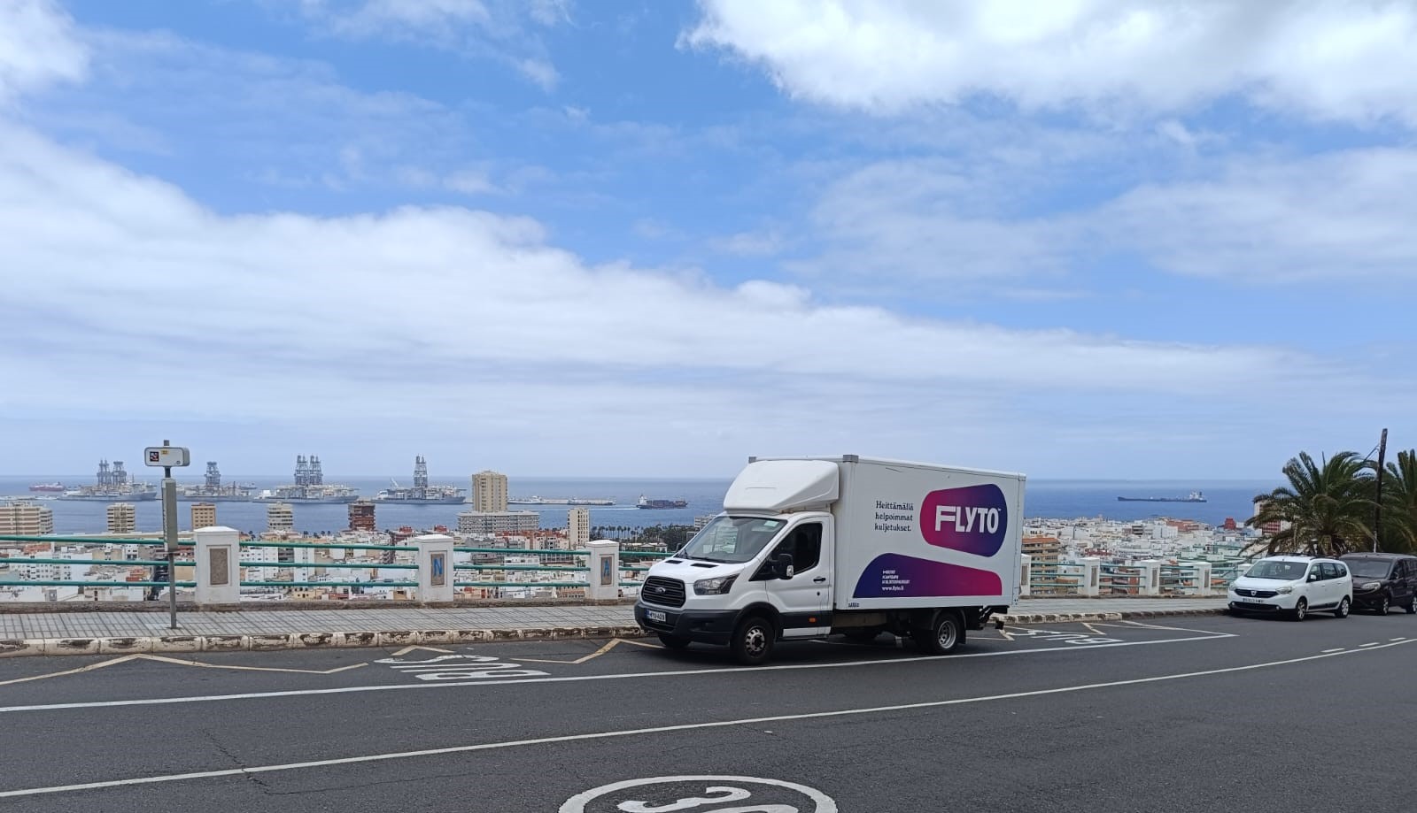 Flyto moving truck driving on coastal road with harbor and ships in background