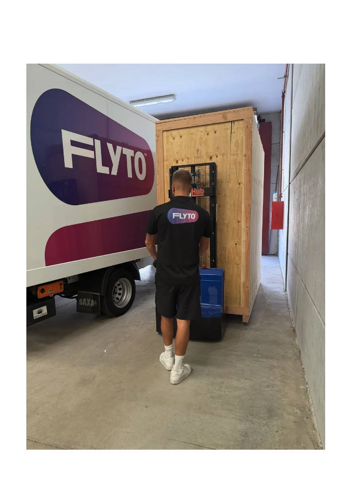 FLYTO moving company worker loading wooden crate near branded delivery truck in warehouse corridor