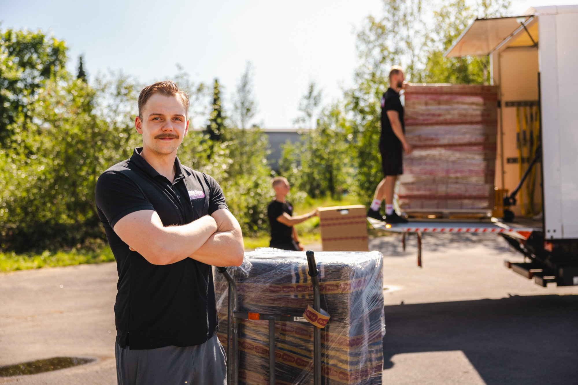 Moving company worker standing beside wrapped pallet with truck being loaded in background