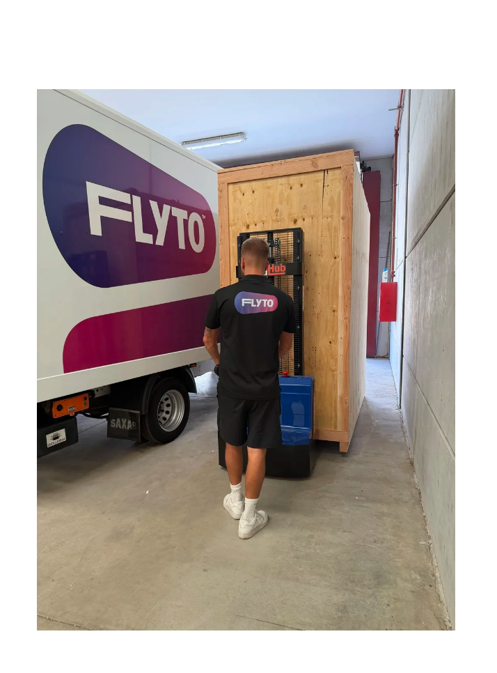 FLYTO moving company worker loading wooden crate near branded delivery truck in warehouse corridor