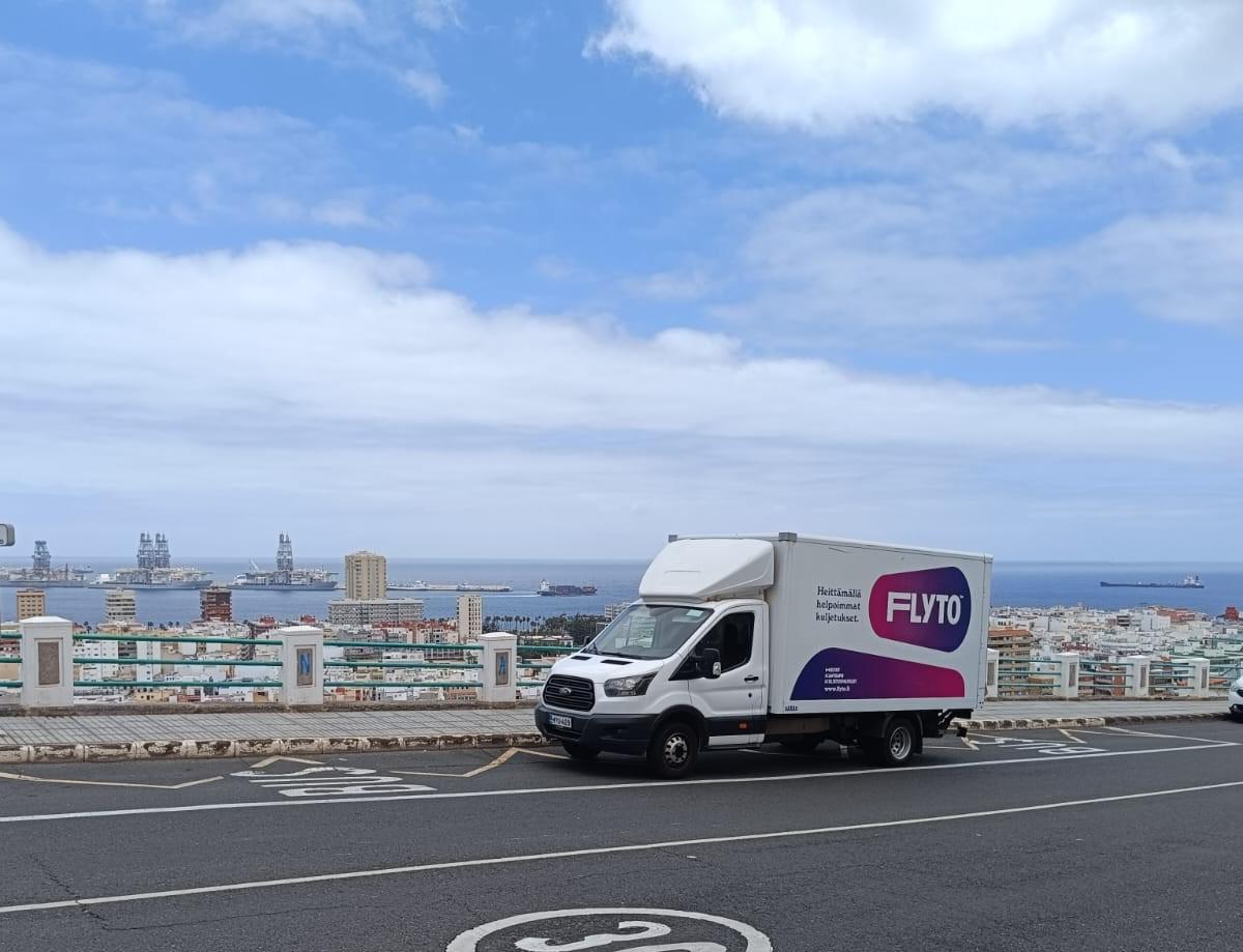 Flyto moving truck on coastal road with harbor and cargo ships in background